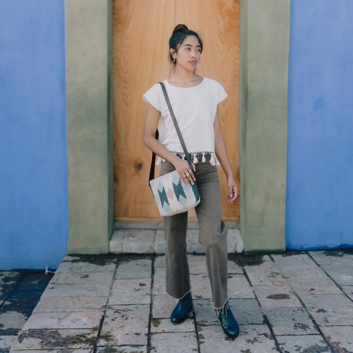 Woman carrying an AMASOUK Espejos Crossbody bag with dark brown leather strap, handwoven wool in blue, heather gray, and ivory, featuring Zapotec patterns. Artisan-made and perfect for everyday use.