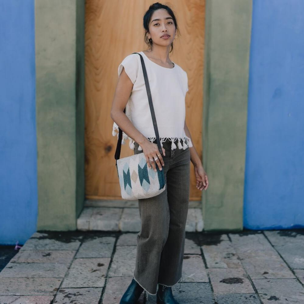 Fashionable woman with an AMASOUK Espejos Crossbody bag, showcasing dark brown leather strap and handwoven wool in blue, heather gray, and ivory. Detailed Zapotec patterns, artisan-crafted for effortless style.