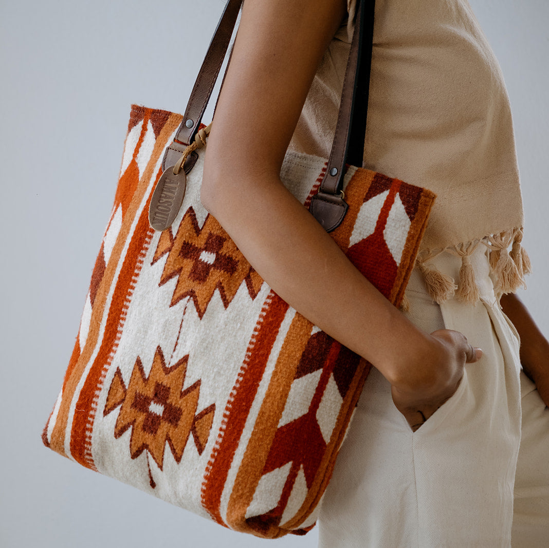 A person wearing a beige dress carries a large woven tote bag with a Southwestern geometric pattern in orange, brown, and cream. The bag has dark brown leather shoulder straps and a small hanging leather tag.