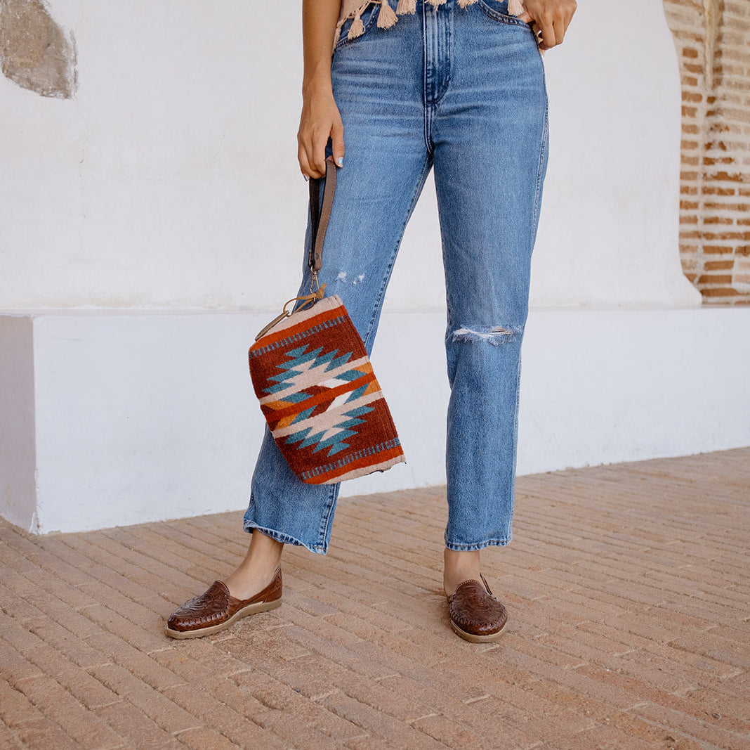 A person wearing light-wash blue cropped jeans and brown leather loafers holds a colorful woven tote bag with a Southwestern geometric pattern featuring red, orange, blue, and white.
