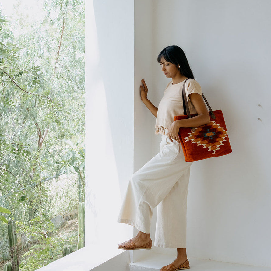 A woman in a cream top and white wide-leg pants stands by a white wall in a bright, modern interior, holding a red woven tote bag with a colorful geometric pattern.
