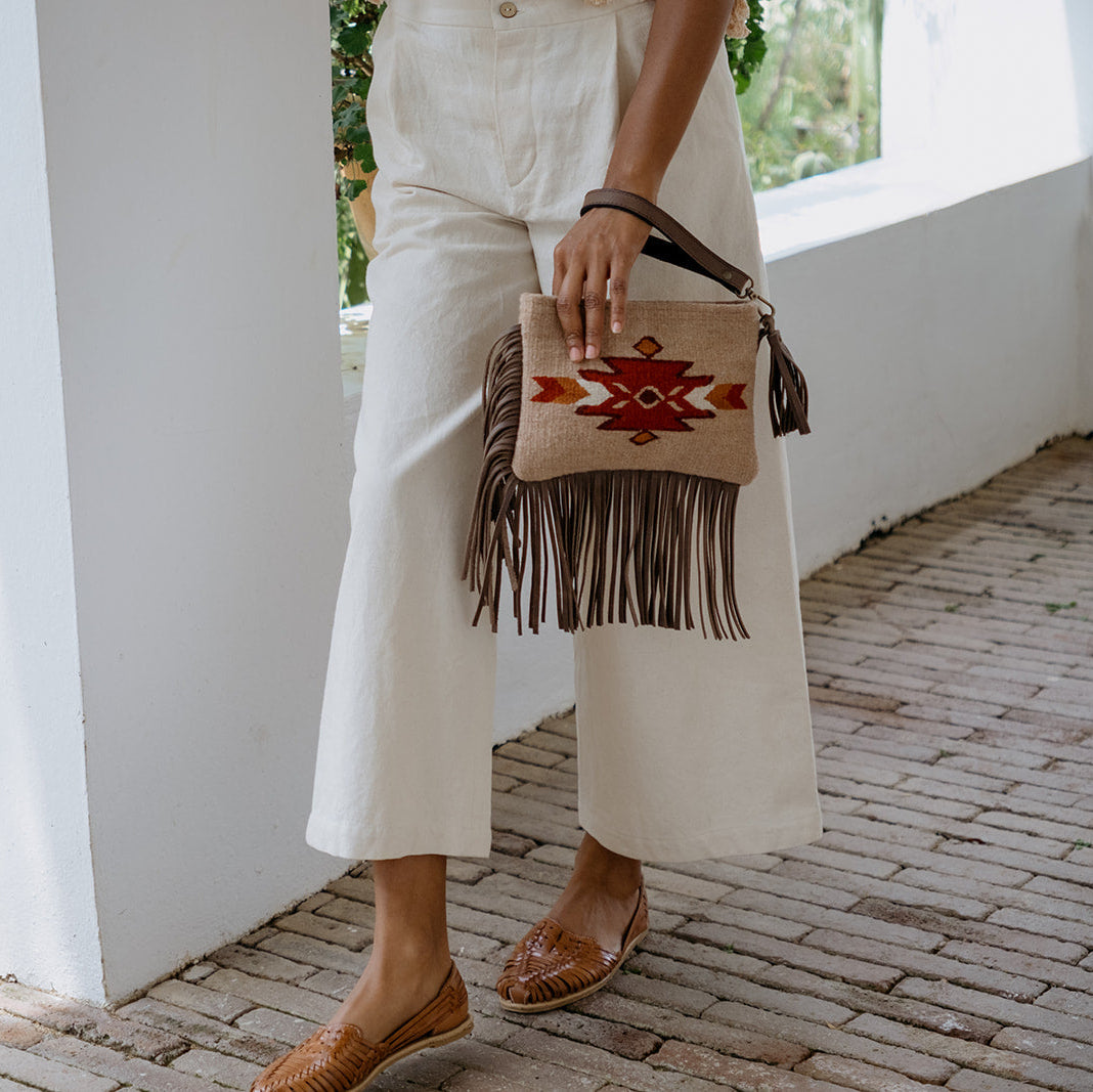 A person wearing white cropped wide-leg pants and brown flat shoes carries a beige woven clutch bag with a red geometric pattern and dark brown fringe, standing on a brick walkway beside white walls.