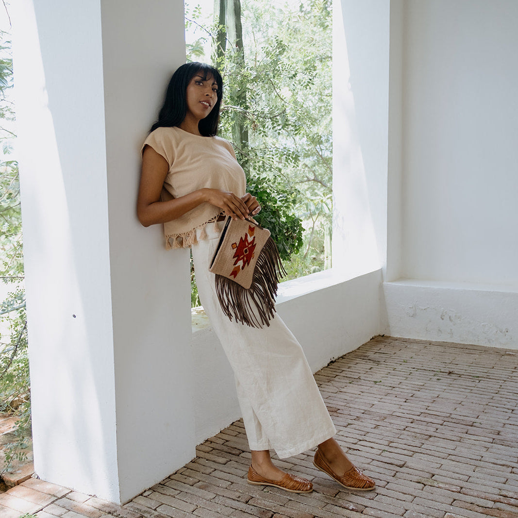 A woman stands barefoot in a sunlit porch setting wearing a beige fringed top and white maxi skirt, holding a brown paper bag with a red graphic print.