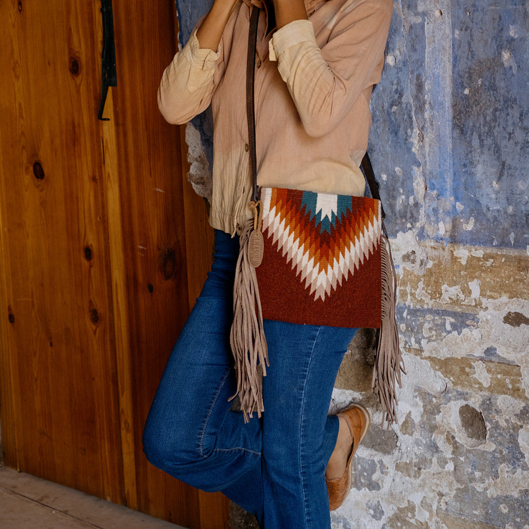 A person wearing a beige cardigan, blue jeans, and tan boots carries a brown woven fringe crossbody bag with a Southwestern-style geometric pattern in red, white, and blue, standing against a rustic blue and brown wall.