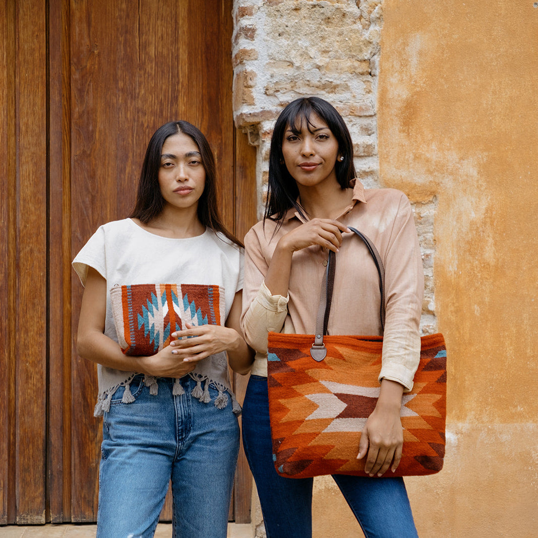 Two women standing outdoors in front of a rustic wooden door and textured wall, each holding a large woven tote bag with a geometric Southwestern-style pattern in orange, red, beige, and blue.