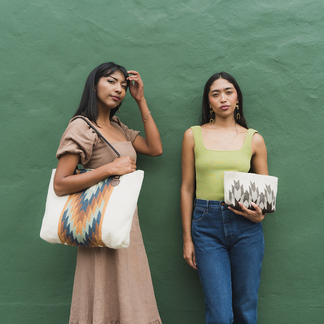 Two women standing against a textured green wall, each holding a patterned tote bag. The woman on the left wears a brown dress and holds a white tote with orange, blue, and black geometric patterns.