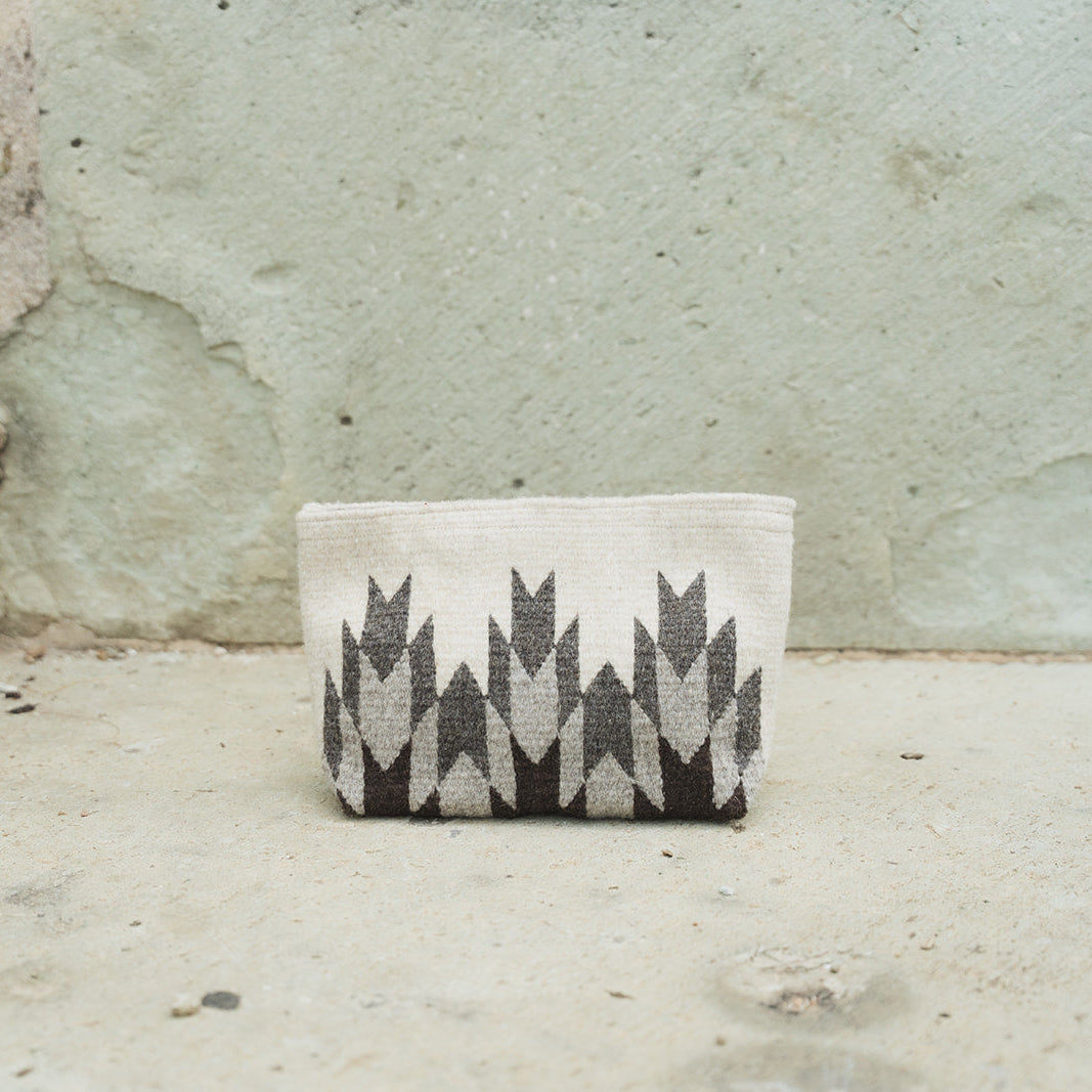 A small woven pouch with a black and off-white geometric pattern, placed on a concrete floor against a textured concrete wall.