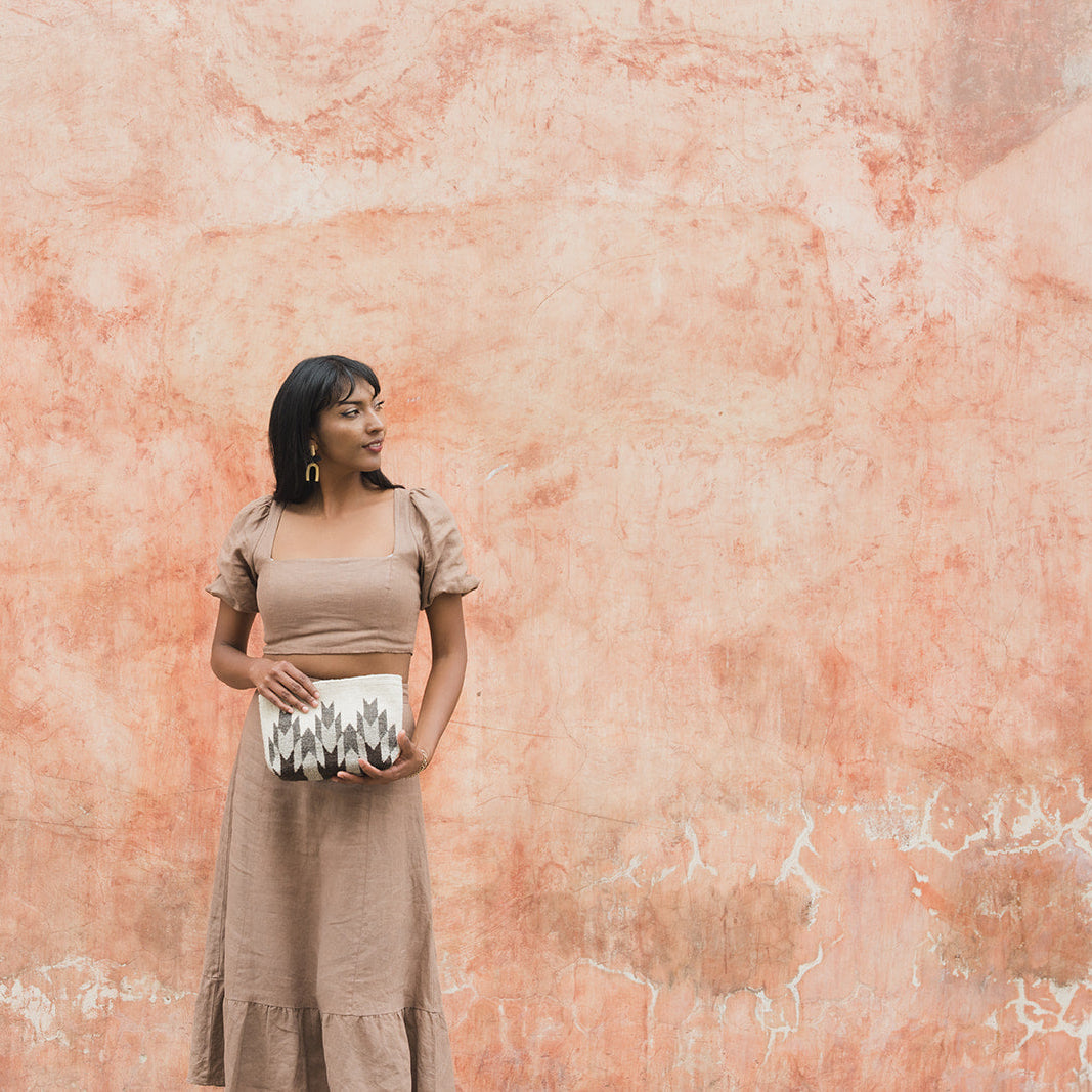 A woman wearing a beige short-sleeve midi dress and holding a black, white, and gray patterned clutch bag, standing in front of a textured terracotta-colored wall.