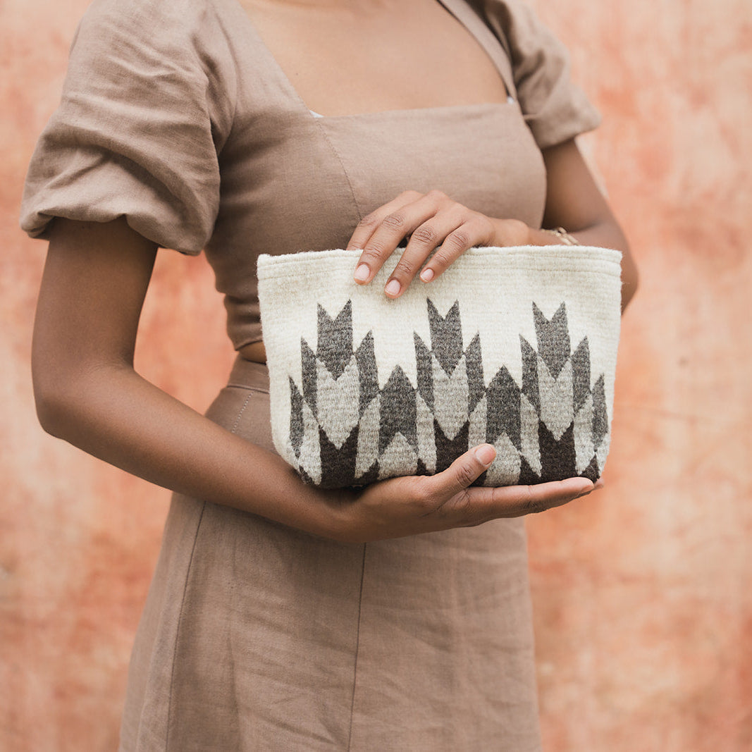 A person in a taupe dress holds a woven clutch bag with a black and white geometric pattern against a textured terracotta background.